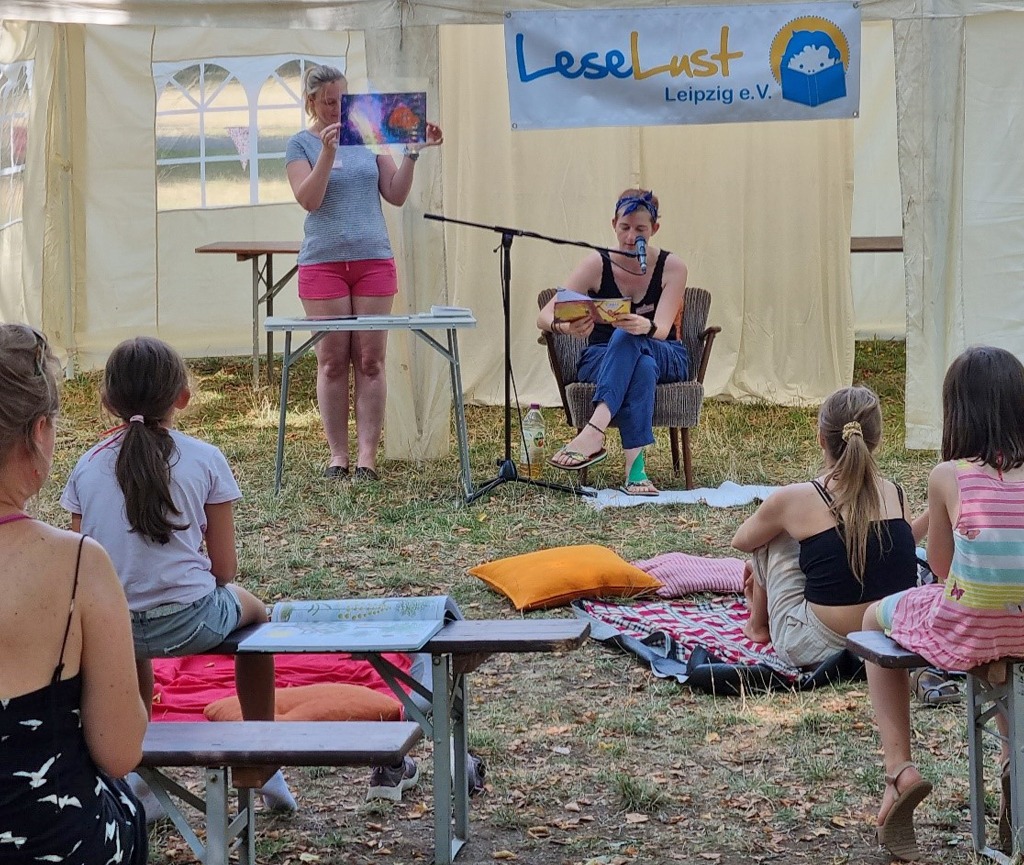 Zwei Vereinsmitglieder lesen auf der Lesebühne. Im Vordergrund sitzen viele Kinder und Eltern und hören interessiert zu.
