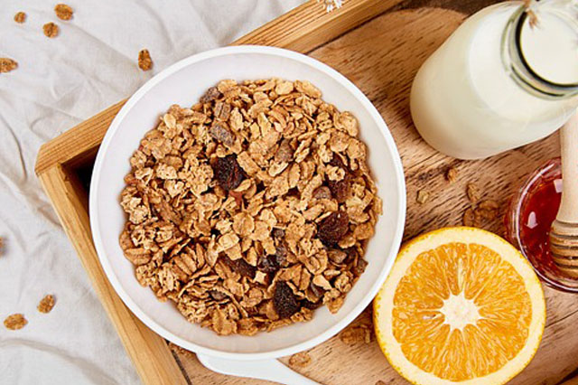 Eine Schüssel mit Frühstücksflocken und eine aufgeschnittene Orange liegen auf einem Holzbrett.