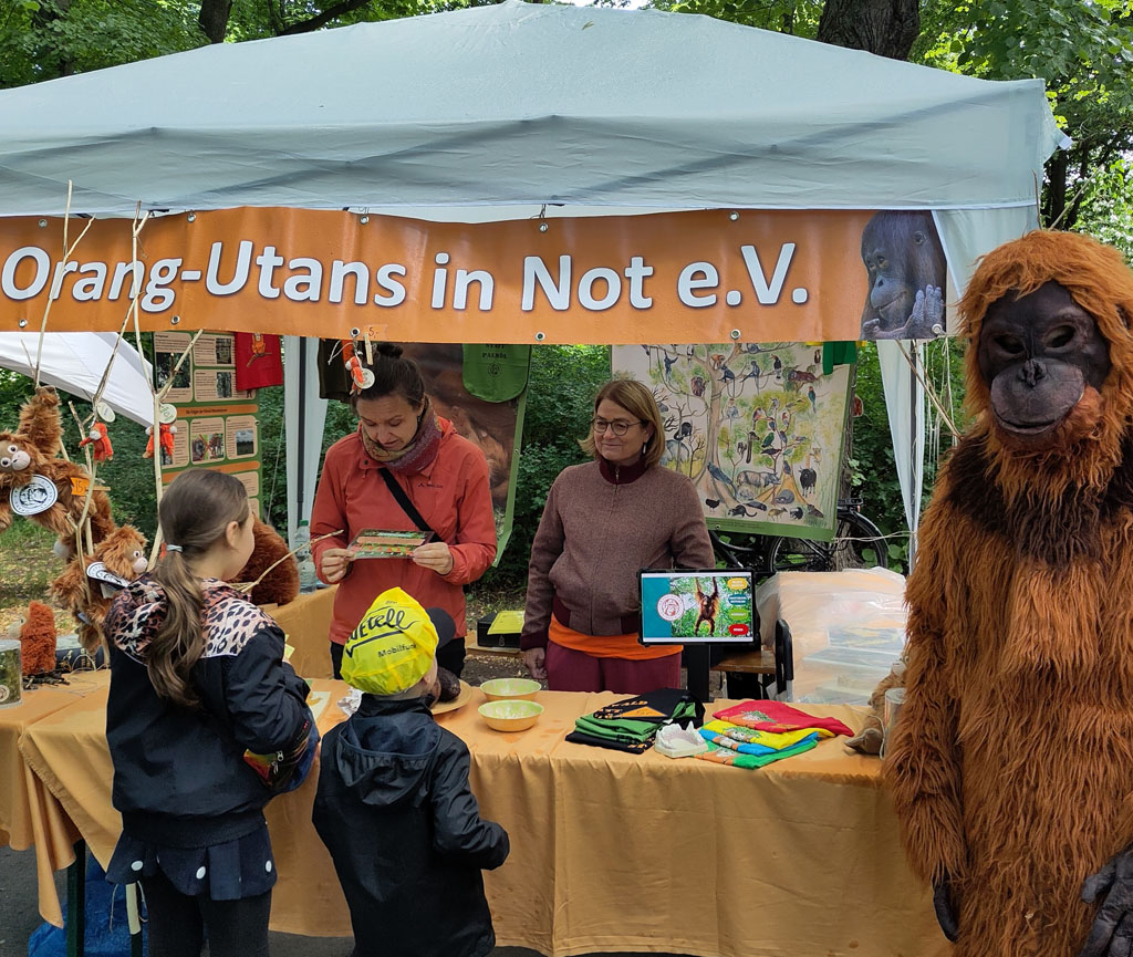 Zwei Kinder stehen vor dem Orang-Utans in Not Infostand. Eine Mitarbeiterin des Vereins spricht mit ihnen.