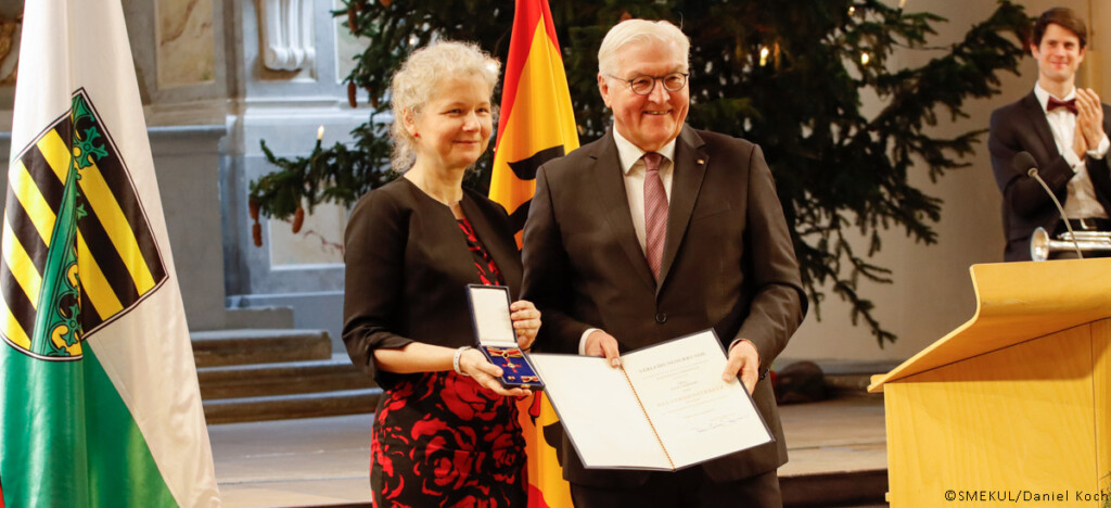 Julia Cissewski steht neben dem Bundespräsidenten Frank-Walter Steinmeier und hält den Bundesverdienstorden in richtung Kamera. Frank-Walter Steinmeier hält die dazugehörige Urkunde in die Kamera.