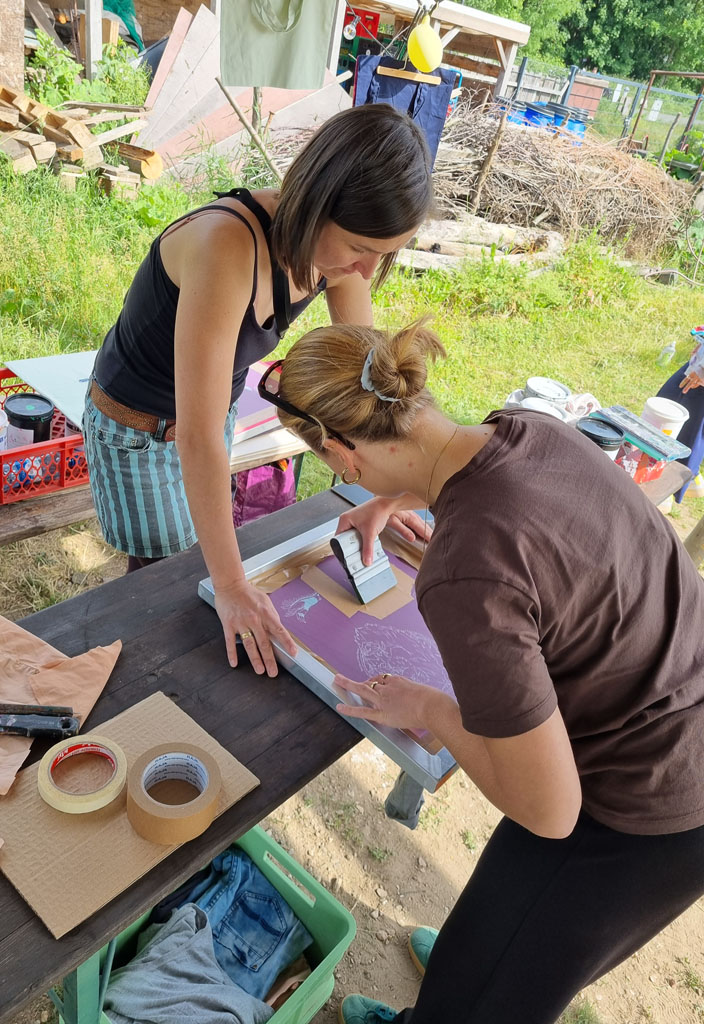 Eine Besucherin an unserem Stand bedruckt ein T-Shirt: Mit einem Siebdruckrakel drückt sie die Farbe durch das Sieb. Eine Mitarbeiterin des Vereins unterstützt sie.