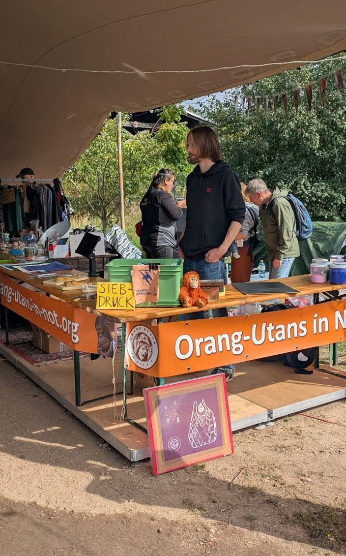 Unter einem großen Sonnensegel ist ein Informationsstand des Orang-Utans in Not e.V. aufgebaut. Dazu gehören zwei große Banner, ein Orang-Utan-Plüschtier und diverse Informationsmaterialien. Im Vordergrund lehnt ein Siebdrucksieb an einer Stange, darüber hängt ein bedrucktes T-Shirt. Auf dem rechten Tisch liegen Farben und andere Materialien für den Siebdruck